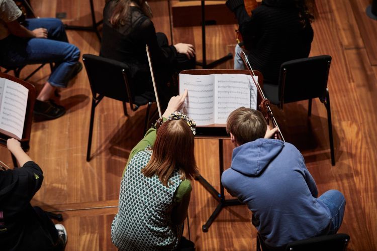 From left to right: A girl wearing a lime green long sleeve top with a spotted blue slip sits next to a boy in a blue jumper. They look at a music book together.